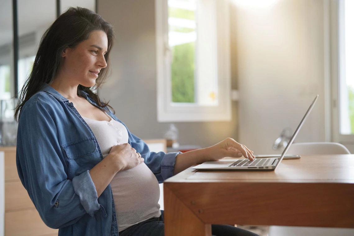 Pregnant woman sitting at a desk, gently holding her belly with one hand while using a laptop with the other.
