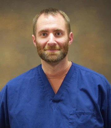 Person wearing blue medical scrubs smiling in front of a brown background.