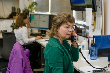 Woman wearing glasses and a green sweater talking on the phone at an office desk, with another person working at a computer in the background.