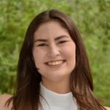 bridget maloney, smiling, wearing a white blouse, had medium length brown hair, greenery in the background